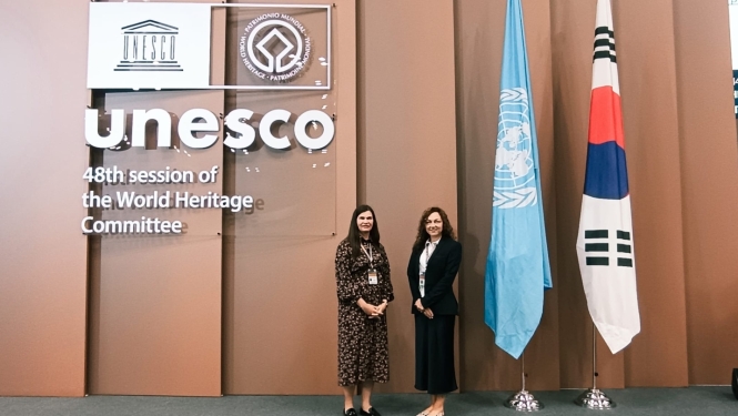 Uz gaišu paneļu fona stāv divas sievietes. Uz fona ir UNESCO logo. Blakus ANO un Korejas Republikas karogs.