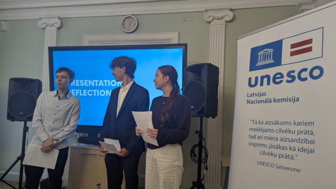 Trīs jaunieši prezentē savas idejas stāvot iekštelpās zila ekrāna priekšā, blakus ir plakāts ar UNESCO LNK logo un tekstu no konstitūcijas