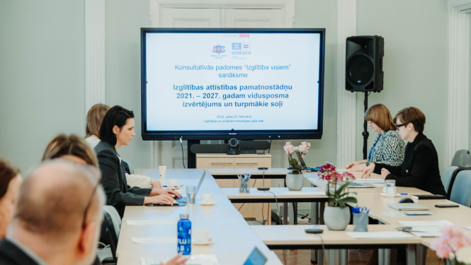 Sanāksmes dalībnieki sēž ap gariem galdiem telpā, kuras centrā ir liels ekrāns ar prezentāciju. Uz ekrāna redzams Izglītības un zinātnes ministrijas un UNESCO logotips, kā arī teksts: "Konsultatīvās padomes 'Izglītība visiem' sanāksme. Izglītības attīstības pamatnostādņu 2021.–2027. gadam vidusposma izvērtējums un turpmākie soļi." Uz galdiem ir ziedi vāzēs, klēpjdatori un ūdens pudeles.