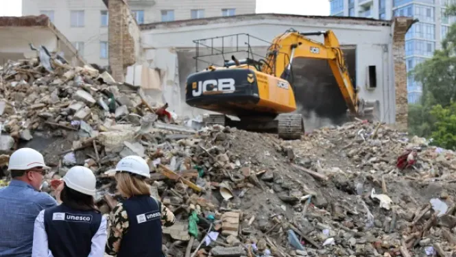 Trīs cilvēki UNESCO vestēs un baltās aizsargķiverēs no mugurpuses vēro ēkas drupas. Fonā dzeltens JCB ekskavators veic būvgružu novākšanas vai nojaukšanas darbus pie pussabrukušas ēkas sienas.