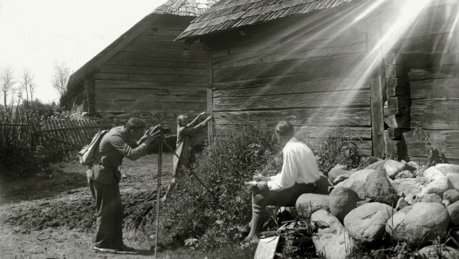 Melnbalta fotogrāfija, saules apspīdēta lauku sēta ar divām guļbaļķu būvēm, ko fotografē vīrietis, kuram asistē viens cits vīrietis un sieviete