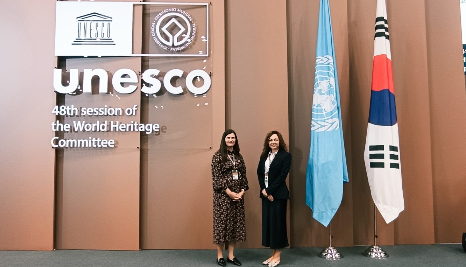 Uz gaišu paneļu fona stāv divas sievietes. Uz fona ir UNESCO logo. Blakus ANO un Korejas Republikas karogs.