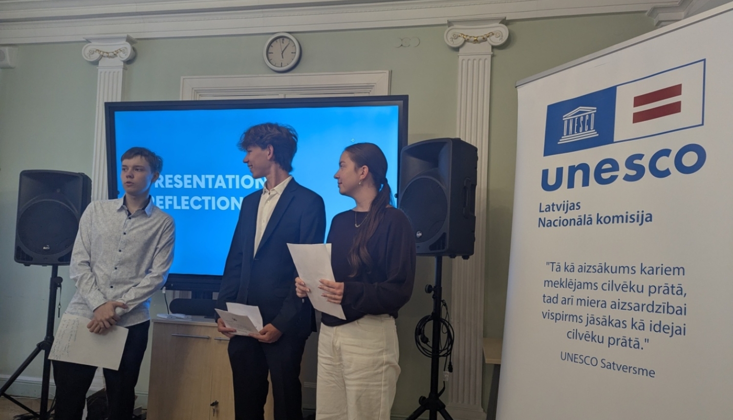 Trīs jaunieši prezentē savas idejas stāvot iekštelpās zila ekrāna priekšā, blakus ir plakāts ar UNESCO LNK logo un tekstu no konstitūcijas