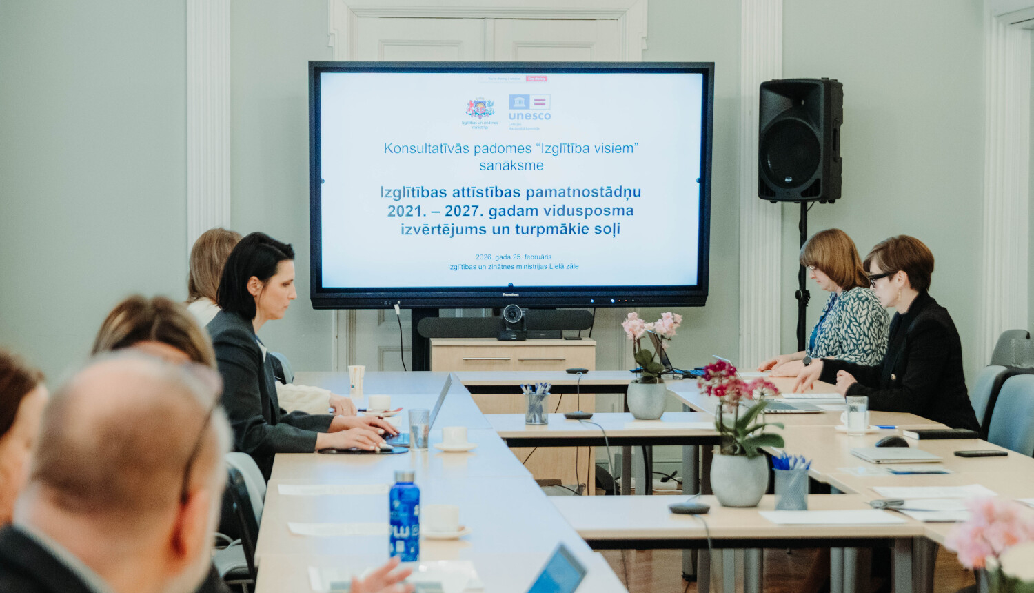 Sanāksmes dalībnieki sēž ap gariem galdiem telpā, kuras centrā ir liels ekrāns ar prezentāciju. Uz ekrāna redzams Izglītības un zinātnes ministrijas un UNESCO logotips, kā arī teksts: "Konsultatīvās padomes 'Izglītība visiem' sanāksme. Izglītības attīstības pamatnostādņu 2021.–2027. gadam vidusposma izvērtējums un turpmākie soļi." Uz galdiem ir ziedi vāzēs, klēpjdatori un ūdens pudeles.