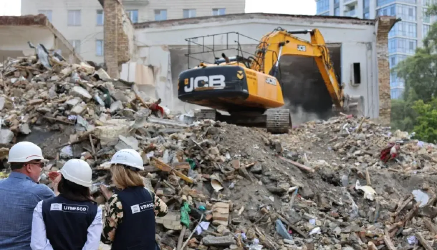 Trīs cilvēki UNESCO vestēs un baltās aizsargķiverēs no mugurpuses vēro ēkas drupas. Fonā dzeltens JCB ekskavators veic būvgružu novākšanas vai nojaukšanas darbus pie pussabrukušas ēkas sienas.