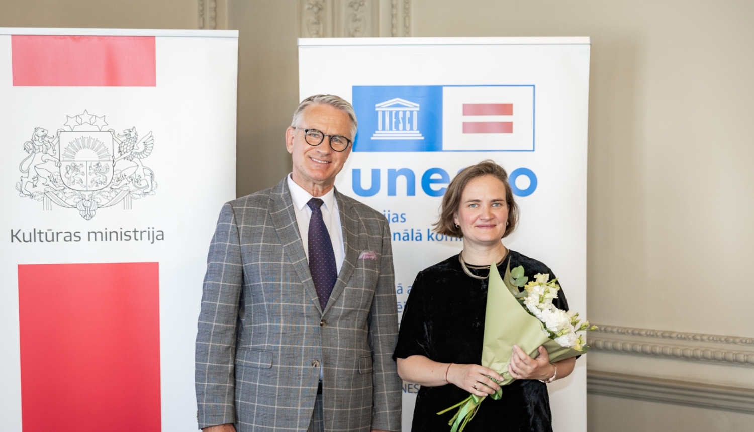 Nauris Puntulis un Dace Resele ar baltu ziedu pušķi, fonā Kultūras ministrijas un UNESCO Latvijas Nacionālās komisijas baneri.