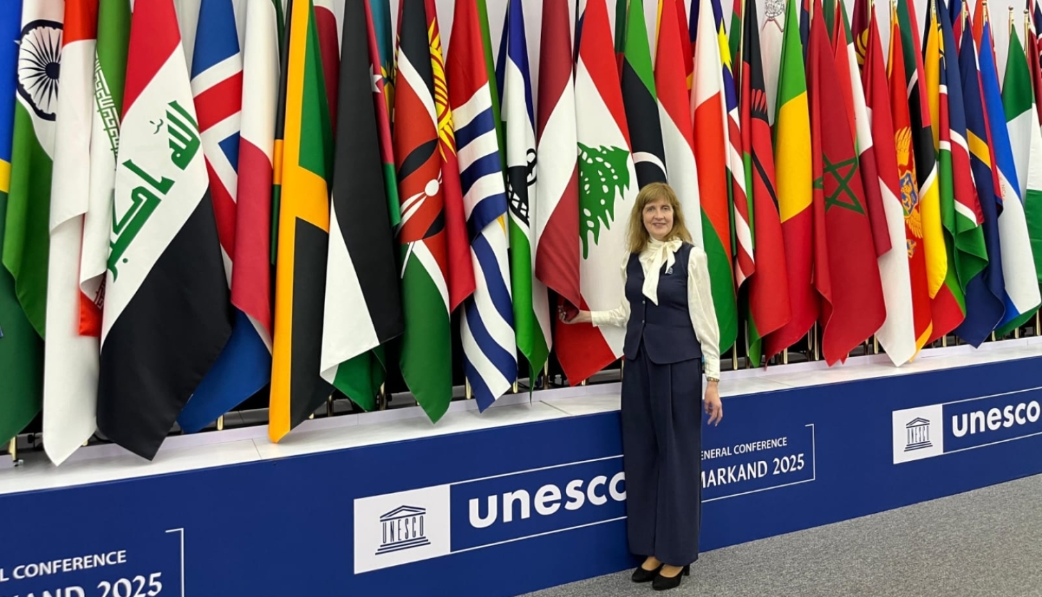 Baiba Moļņika stāv pie dažādu valstu karogiem un UNESCO baneriem