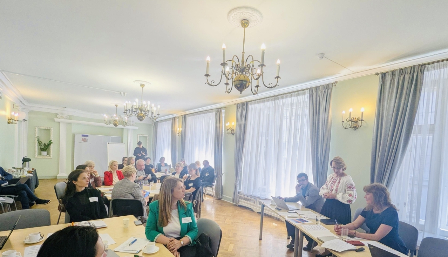 Sanāksmes dalībnieki sēž pie galdiem plašā telpā, runā izglītības un zinātnes ministre Dace Melbārde.