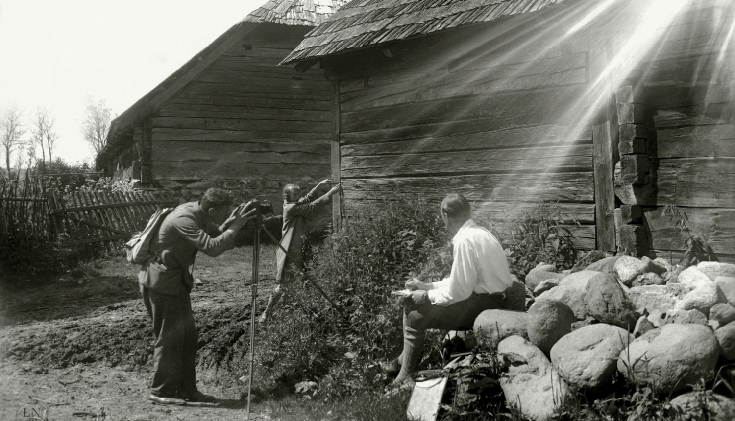 Melnbalta fotogrāfija, saules apspīdēta lauku sēta ar divām guļbaļķu būvēm, ko fotografē vīrietis, kuram asistē viens cits vīrietis un sieviete