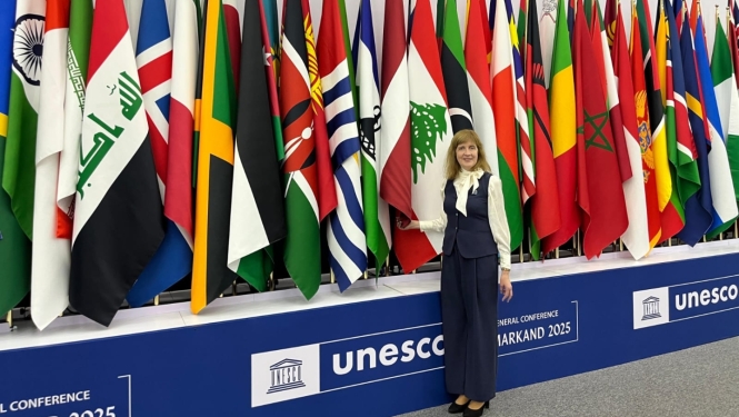 Baiba Moļņika stāv pie dažādu valstu karogiem un UNESCO baneriem