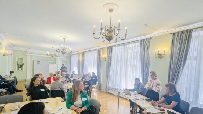 Sanāksmes dalībnieki sēž pie galdiem plašā telpā, runā izglītības un zinātnes ministre Dace Melbārde.
