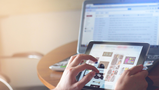 Persona ar rokām izmanto planšetdatoru uz galda, kamēr fonā redzams atvērts klēpjdators ar e-pasta logu.