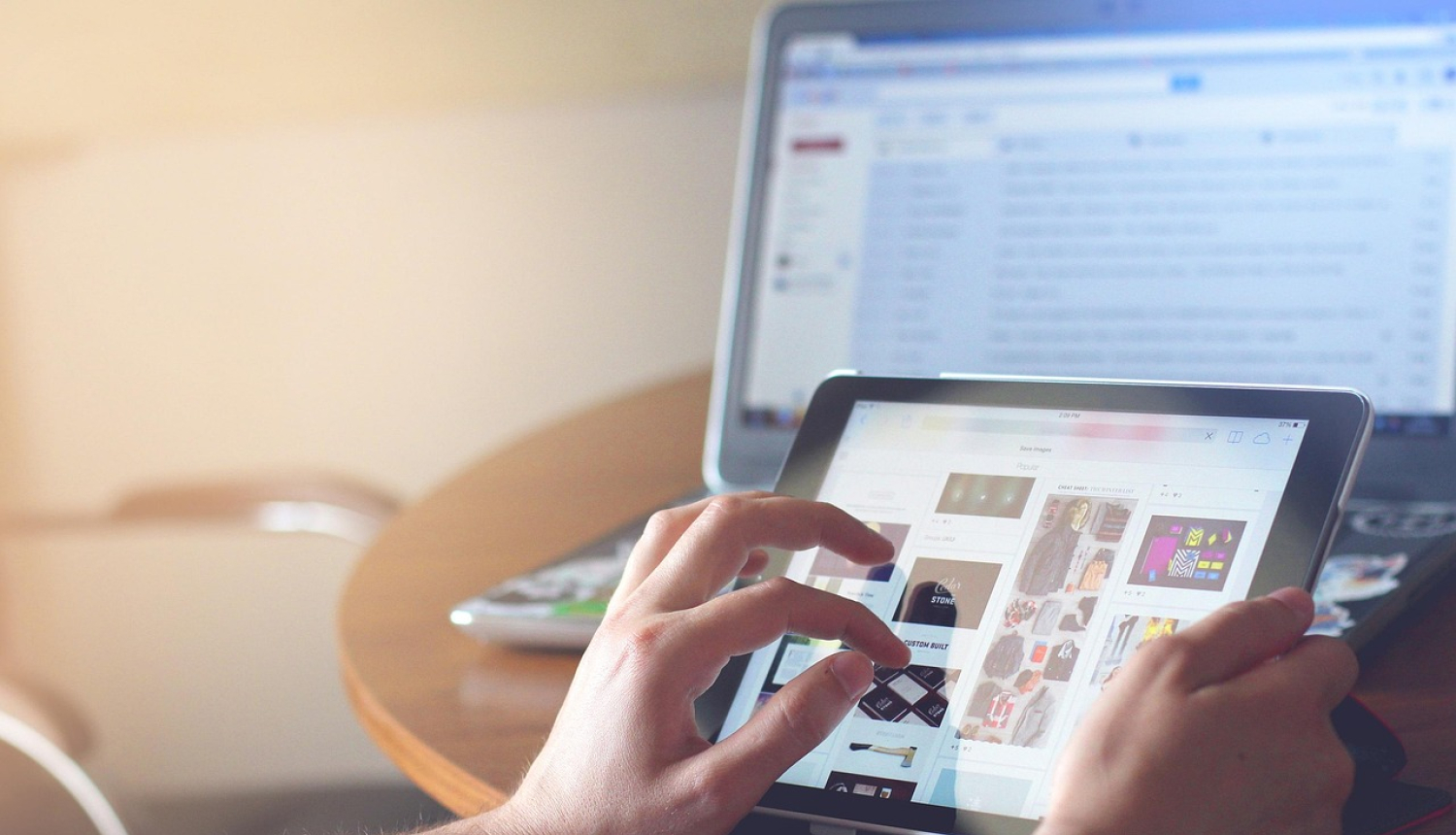 Persona ar rokām izmanto planšetdatoru uz galda, kamēr fonā redzams atvērts klēpjdators ar e-pasta logu.